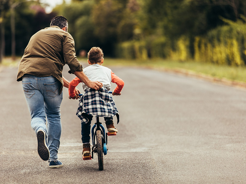 Rear view of a boy riding a bicycle while his father runs along holding the kid. Father teaching his son to ride a bicycle.